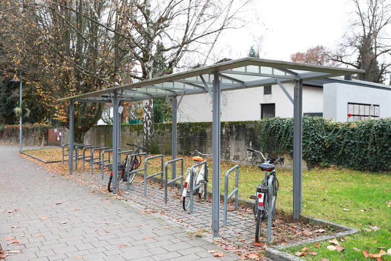 Es sind überdachte Fahrradabstellanlagen zu stehen. Darunter stehen drei Fahrräder. Hinter der Überdachung ist eine Wiese und ein Haus mit Hecke.
