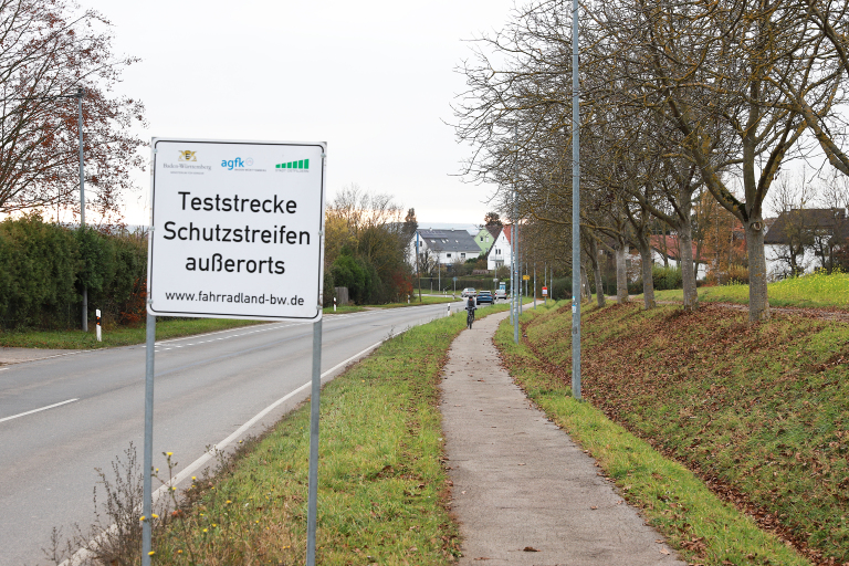 Schild Teststrecke Schutzstreifen außerorts neben einer Straße. Neben der Straße befindet sich ein Grünstreifen und daneben ein Fußgängerweg. 
