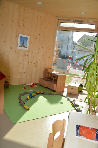 Blick in den Baubereich. Auf dem grünen Spielteppich sind Schienen für eine Spielzeugeisenbahn verlegt. Hier und da stehen Regale mit verschiedenen Spielsachen.