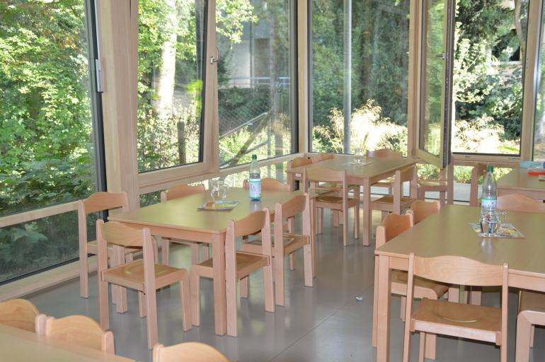 Blick in einen Raum mit großen Fenstern, die einen Ausblick auf eine grüne Umgebung ermöglichen. In diesem Raum stehen Tische mit Stühlen, auf denen weitere Stühle platziert sind.