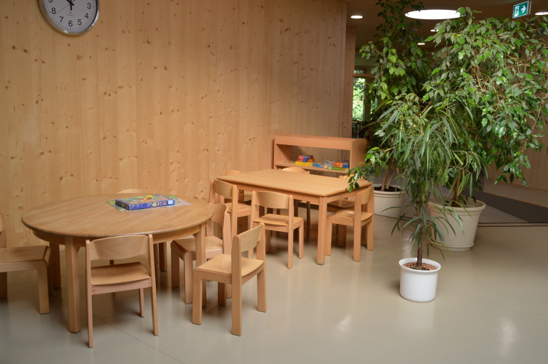 Blick  auf einen Platz mit zwei Tischen und Stühlen. Auf den Tischen liegen Spiele, die aus dem Regal dahinter genommen werden können. Auf der rechten Seite grenzen zwei große Pflanzen den Bereich ab.