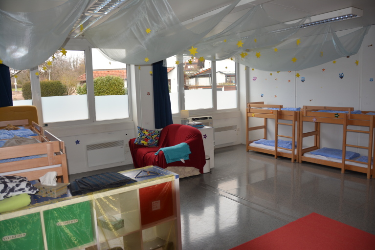 Ein Blick in einen Raum, in dem Kinderbetten und ein roter Sessel zu sehen sind. An der Decke hängt ein weißer, geraffter Stoff mit gelben Sternen.