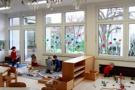 Blick in den Baubereich, der durch ein Regal in zwei Bereiche geteilt ist. In beiden Bereichen sitzen Kinder auf dem Boden und bauen etwas. Im Hintergrund sind Fenster zu erkennen, die mit bunten Blumen bemalt sind.