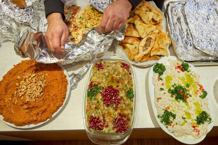 Bei dem Bild handelt es sich um ein Foto. Auf dem Foto sieht man wie mehrere syrische Gerichte von einem Paar Hände auf einem Tisch angerichtet werden.