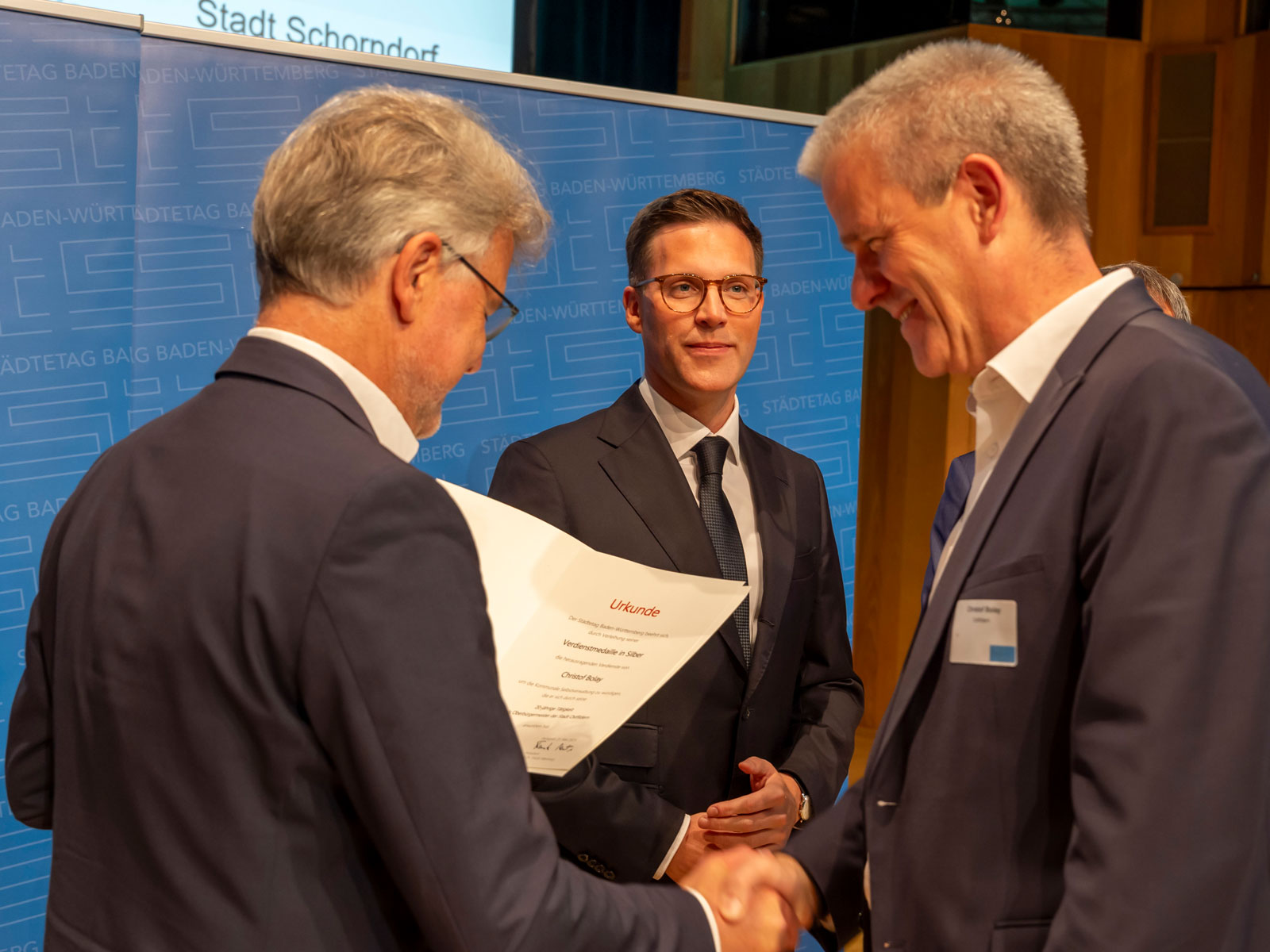 Frank Mentrup, Oberbürgermeister der Stadt Karlsruhe und Präsident des Städtetags Baden-Württemberg, die Ehrungsurkunde an Christof Bolay. In der Bildmitte ist Manuel Hagel, Innenminister und stellvertretender Ministerpräsident des Landes Baden-Württemberg, zu sehen